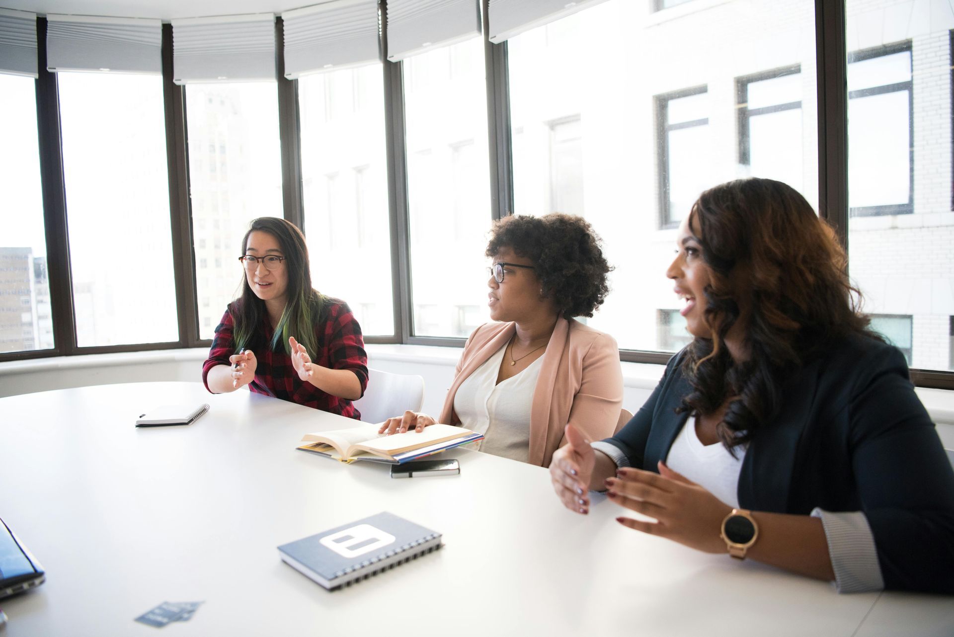 Three women in a meeting around a white table in a bright office; one speaks animatedly.