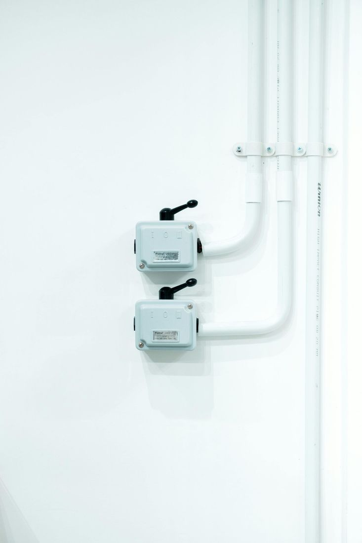 Two white wall-mounted electrical boxes with black switches and pipes on a white wall