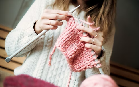 A woman is knitting a pink scarf with knitting needles.