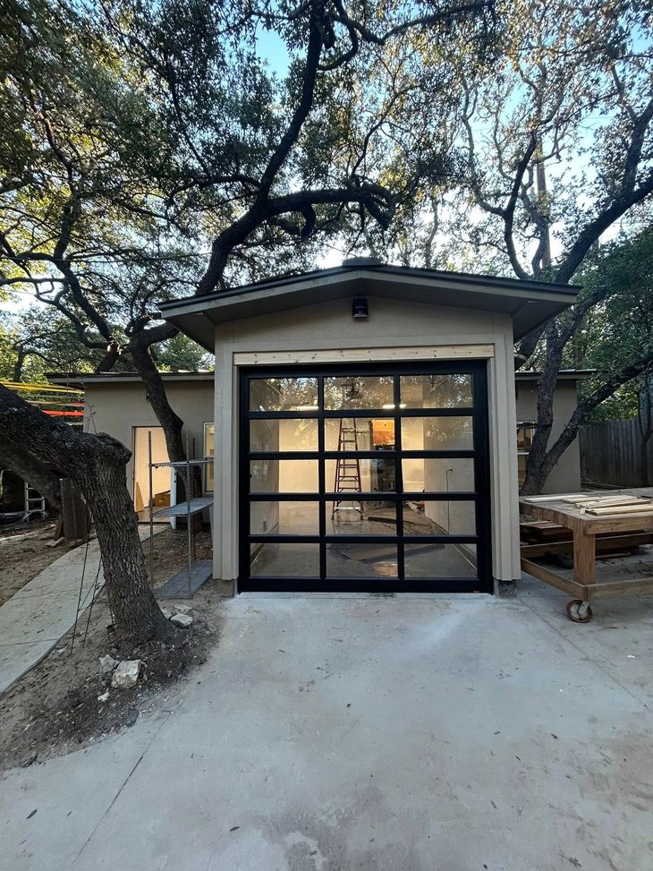Small garage with a black-framed glass door, surrounded by trees on a concrete driveway.