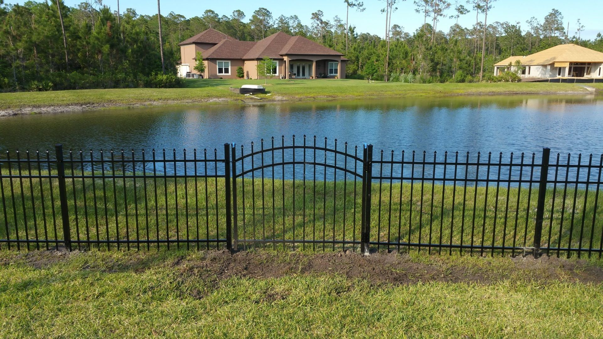 Black metal fence with arched gate bordering a lake, two houses in the background.