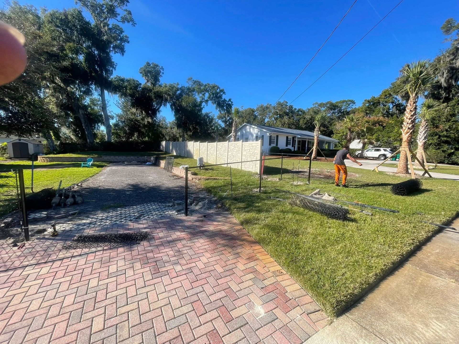 Construction of a driveway with brick pavers, a worker stands nearby, on a sunny day.