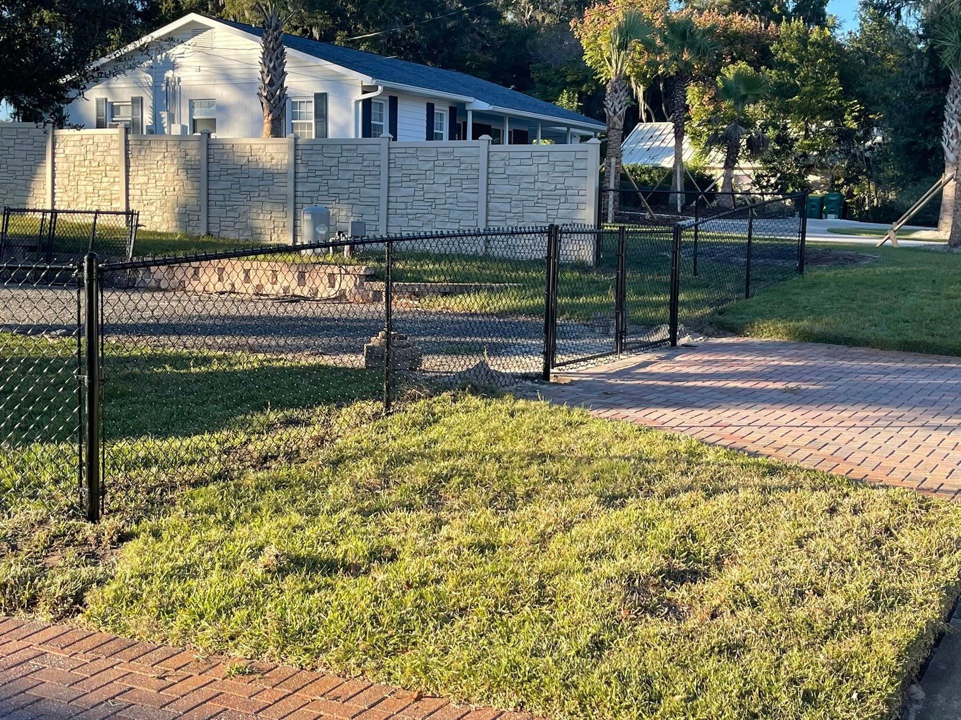 Black chain-link fence on a green lawn in front of a light-colored house. Brick pathway and trees in the background.