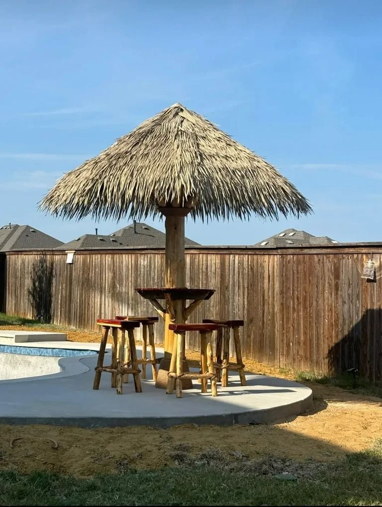Tiki bar with thatched roof and stools, set on a concrete pad near a pool and wooden fence.