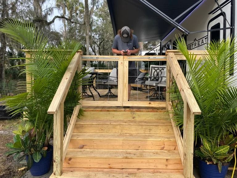 Wooden steps leading to a deck beside an RV; a man stands on the deck. Potted plants flank the stairs.