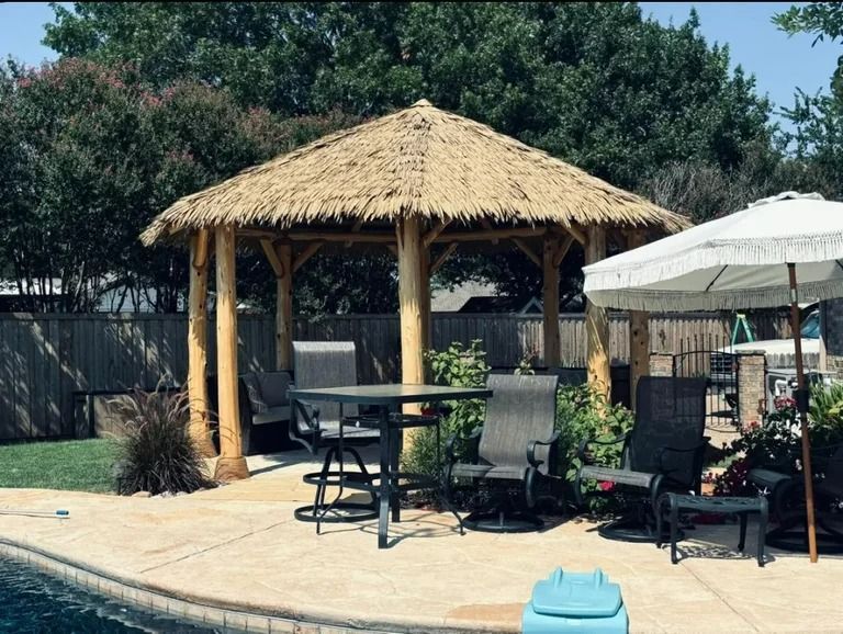 Tiki-style gazebo with a thatched roof, gray chairs, and a table beside a pool in a backyard setting.