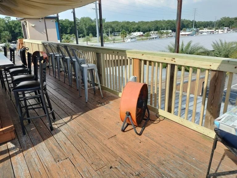Outdoor bar with seating, view of water. Orange fan, wood deck, light wood railing, overcast.