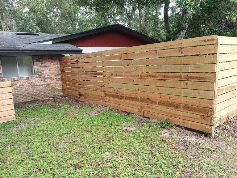 Wooden horizontal slat fence in a grassy yard, partially obscuring a brick house.