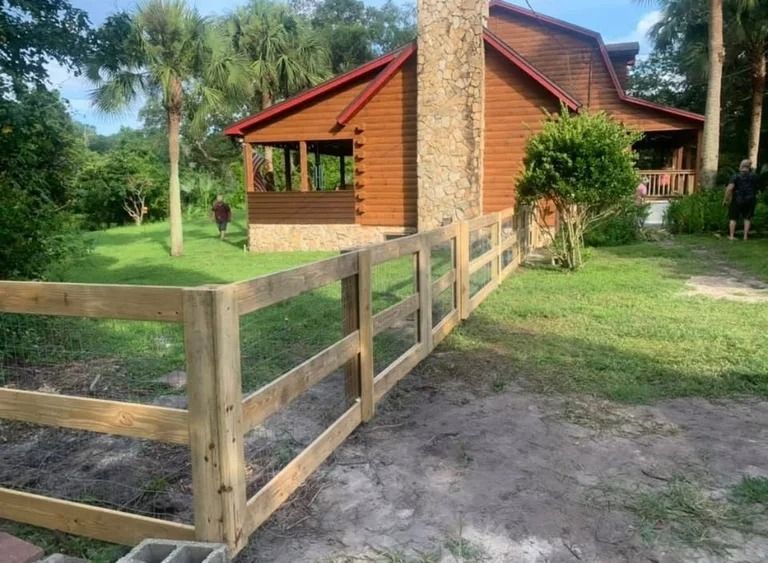 Wooden fence in front of a brown house with stone chimney and red roof; green lawn and trees in background.
