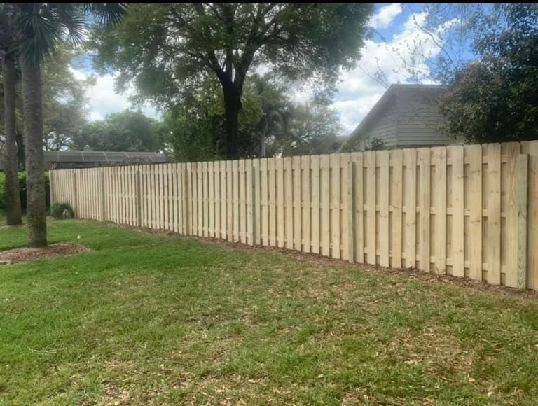 Wooden fence along green grass in front of a house under a cloudy sky.