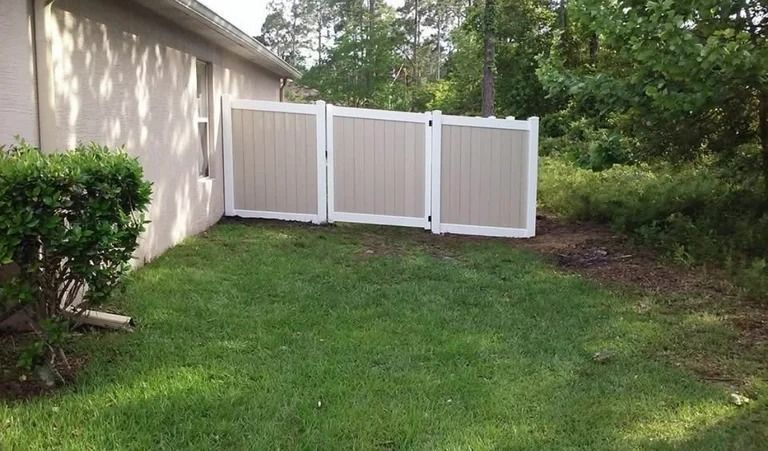 Tan and white fence enclosing a small grassy area next to a beige house, with trees in the background.