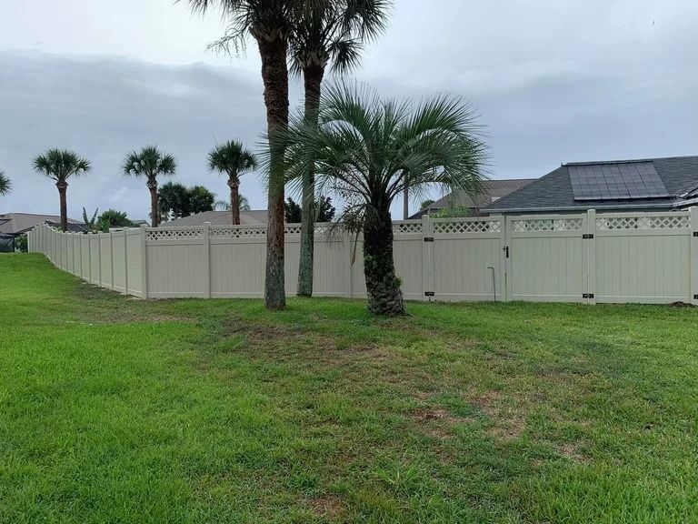 A beige vinyl fence encloses a green lawn, with palm trees and houses in the background under a cloudy sky.