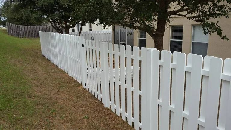 White picket fence along a grassy yard, beside a tan building with windows and a large tree.