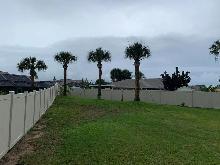 Backyard with white fence, palm trees, and overcast sky. Green grass in foreground.