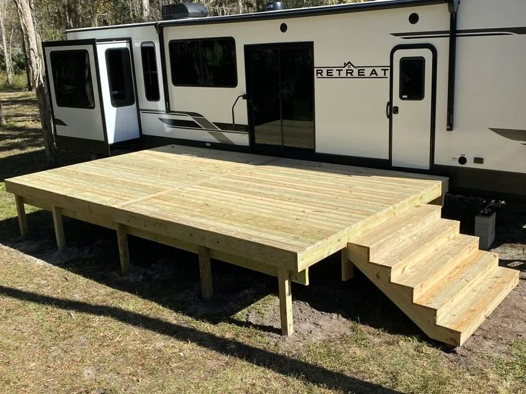 Wooden deck with stairs next to a large RV. The deck is newly built and sits on grass.