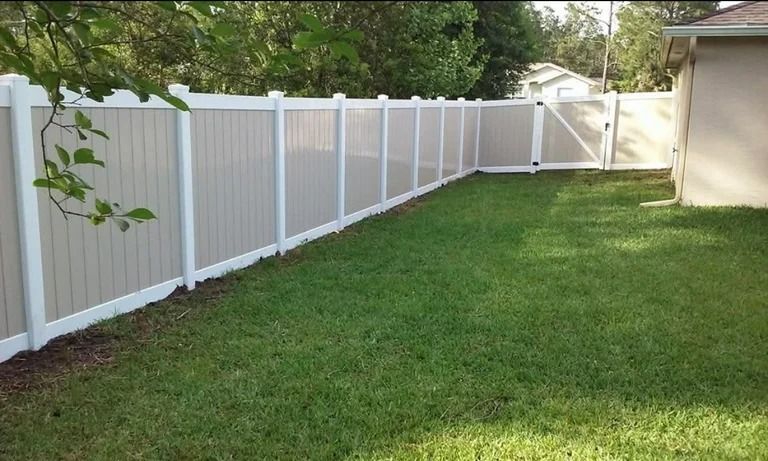 Tan and white vinyl fence enclosing a green grassy yard with a gate, next to a beige building.