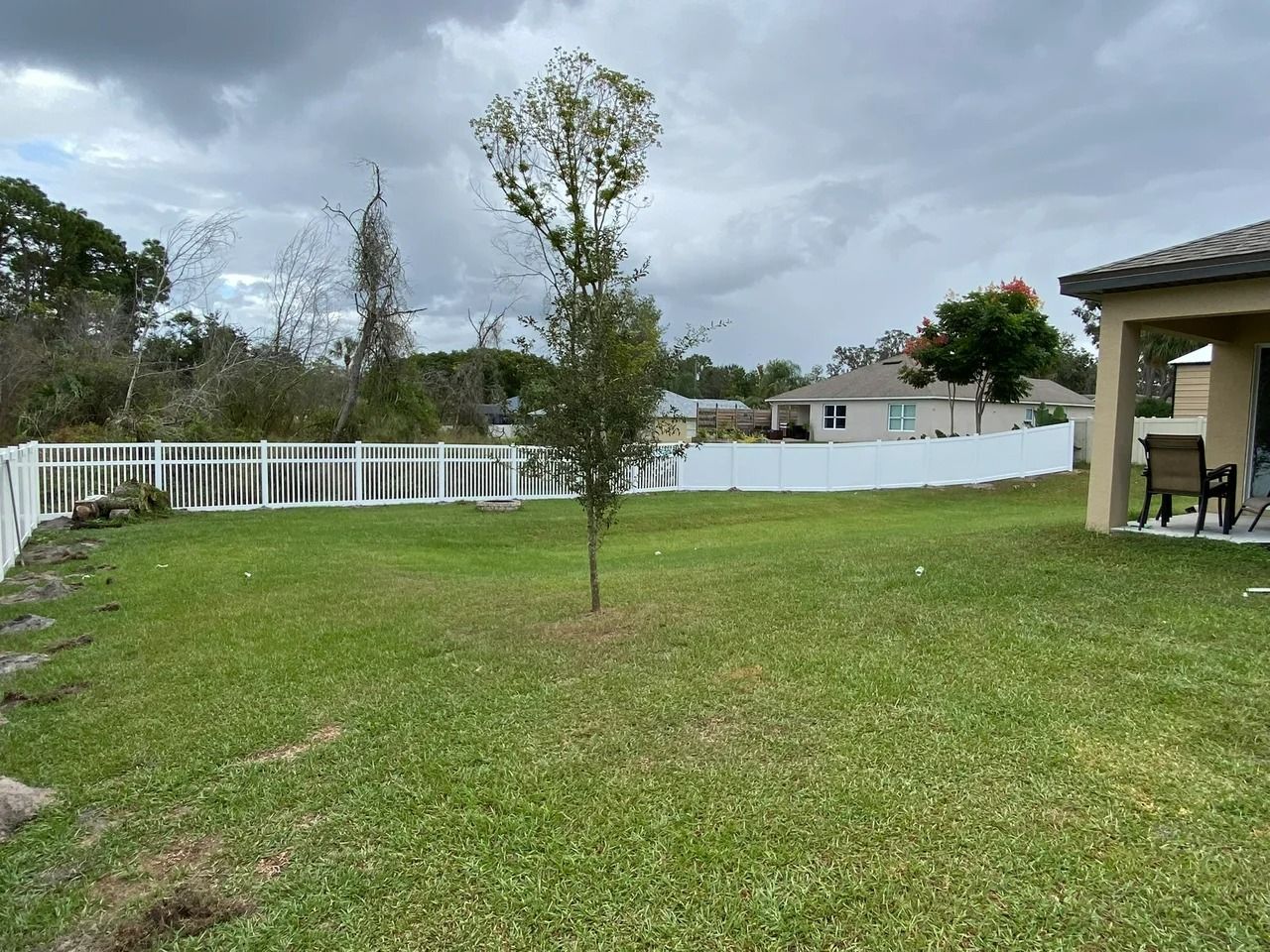 A backyard with green grass, a white picket fence, a small tree, and a home under a cloudy sky.