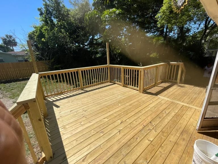 Wooden deck with railings in a backyard setting, surrounded by trees and a fence.