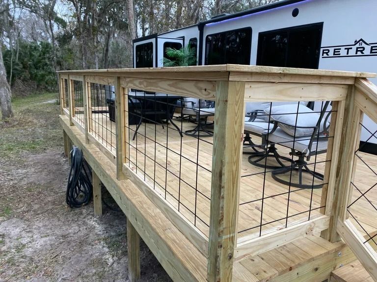 Wooden deck with black metal railing in front of a recreational vehicle. Outdoor seating is visible.