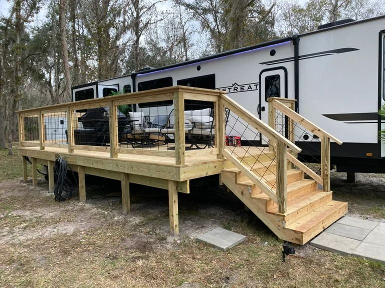 Wooden deck with stairs next to a white RV, set in a grassy outdoor area.