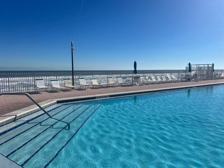 A large swimming pool surrounded by chairs and umbrellas with a view of the ocean.