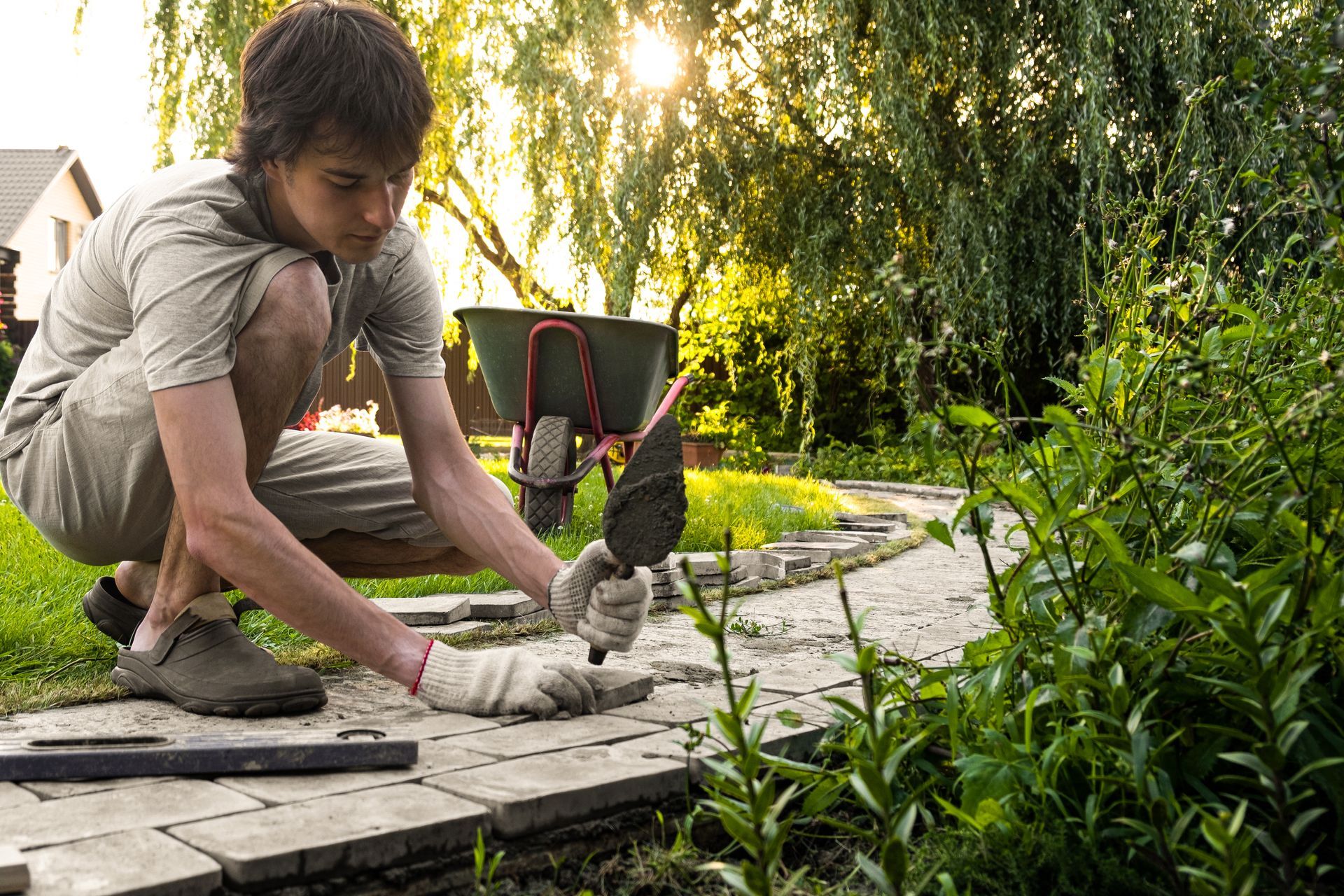 A man is kneeling down in a garden with a shovel and a wheelbarrow.