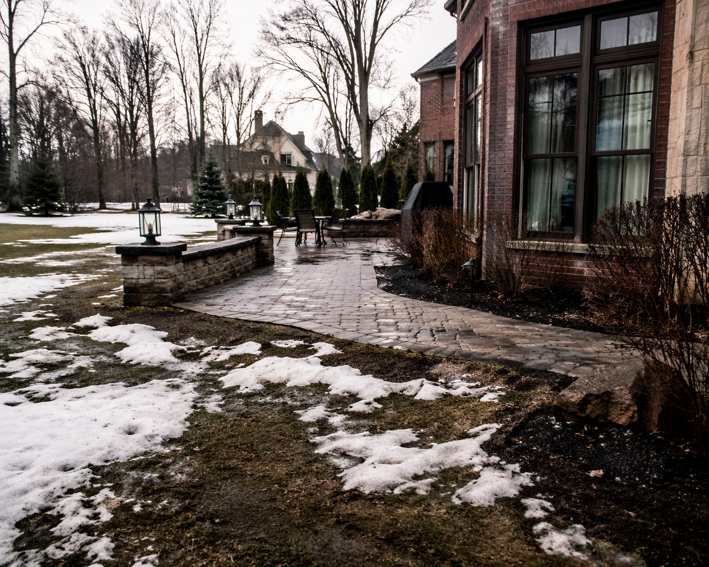 Snowy yard beside brick houses with a stone patio and leafless trees