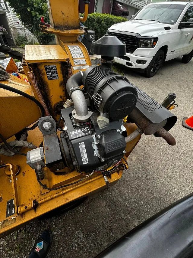 A yellow snow blower is parked on the side of the road next to a white truck.