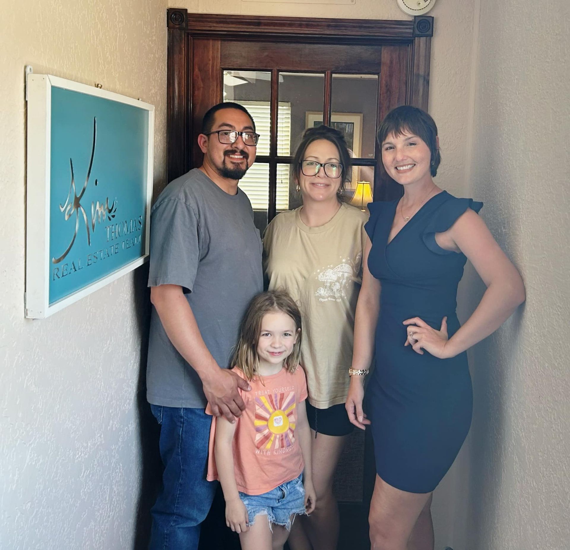 Family and woman in a navy dress pose in a doorway with a sign on the wall; they appear happy.