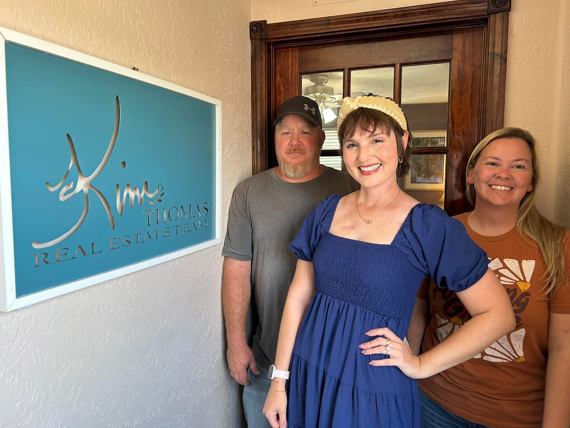 Three people outside a real estate office with a turquoise sign. The woman in blue smiles.
