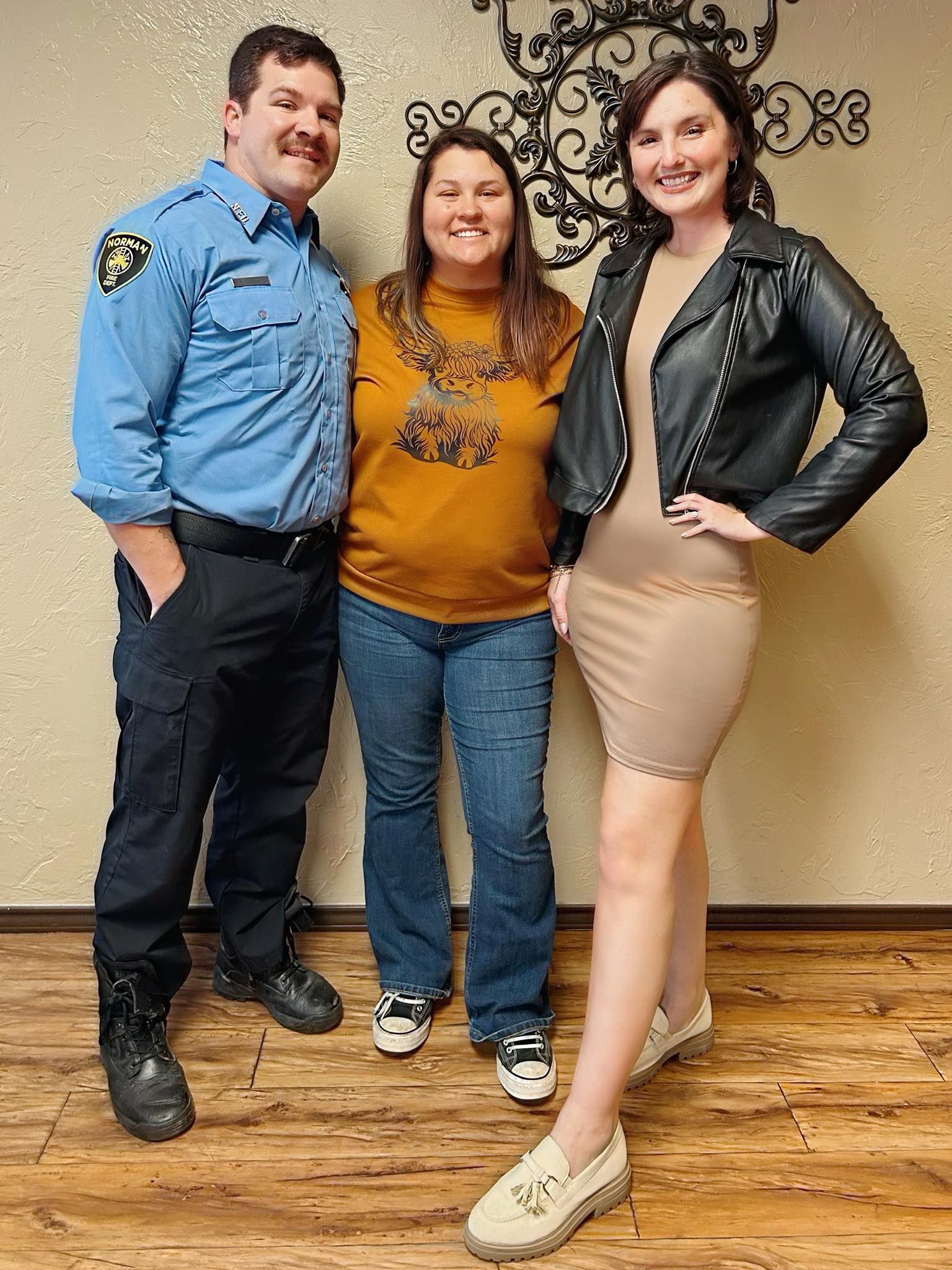 Three people posing: a man in a blue uniform, a woman in an orange shirt and jeans, and a woman in a black jacket and dress.