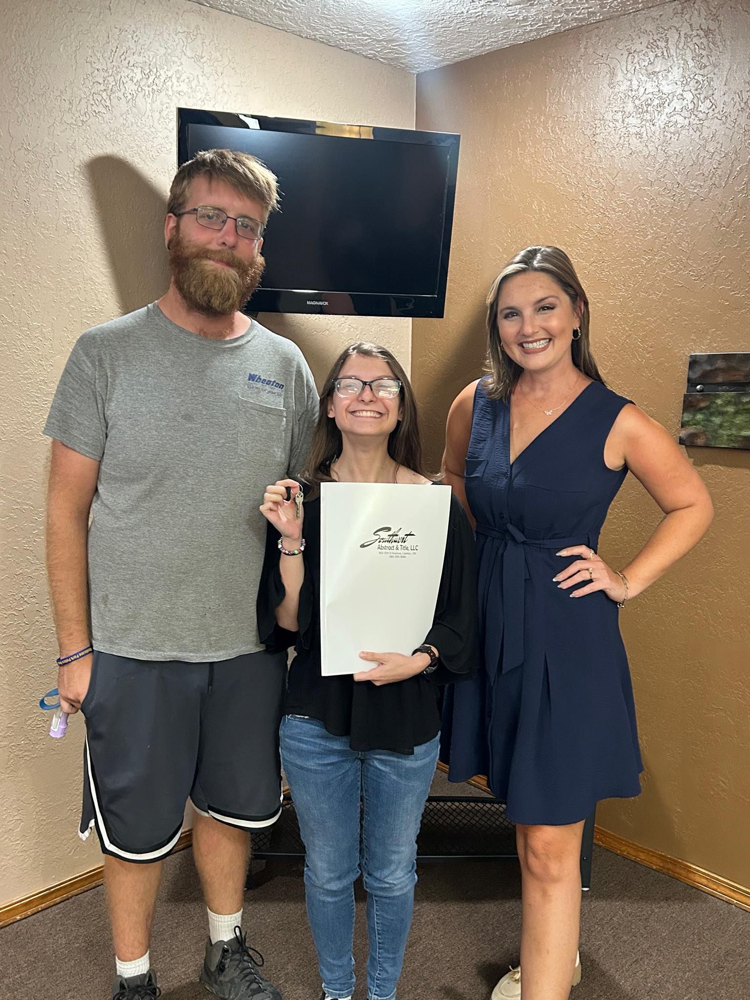 Three people pose indoors: a man, a person holding a document, and a woman. They smile, with a TV behind them.