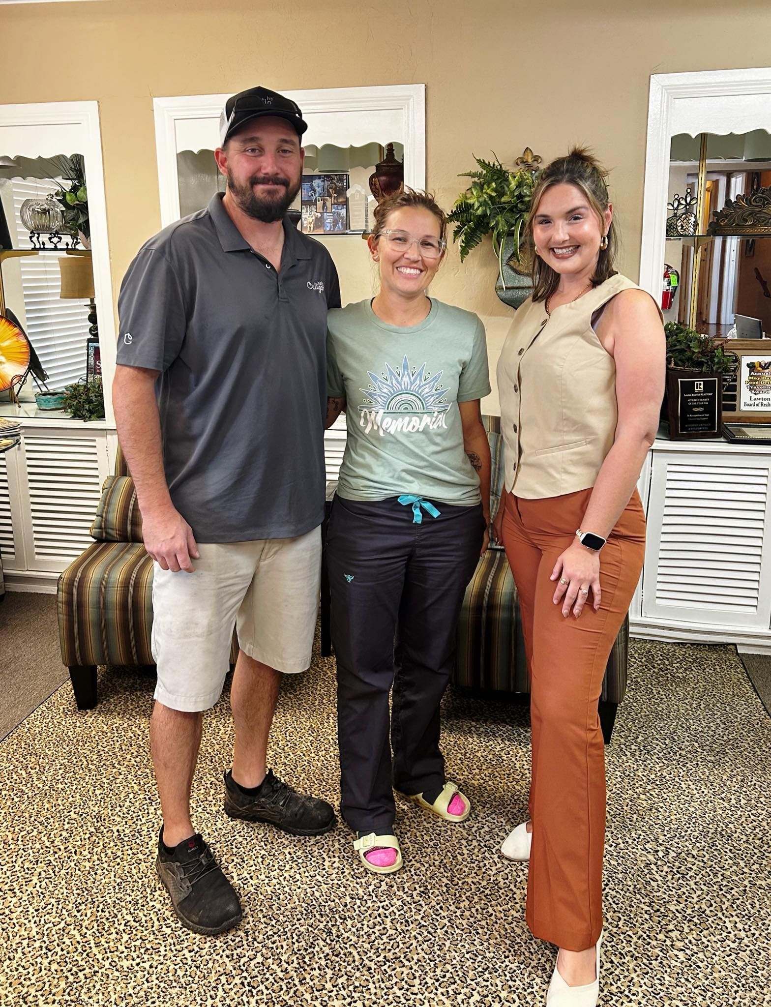 Three people smiling, posing indoors. Man in shorts, cap. Woman in scrubs. Another woman in pants, top.