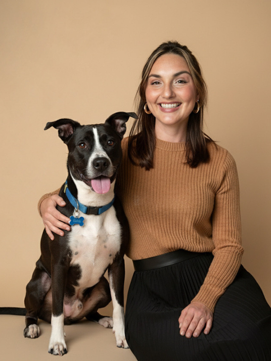 Woman smiles with a black and white dog; both sit against a tan backdrop.