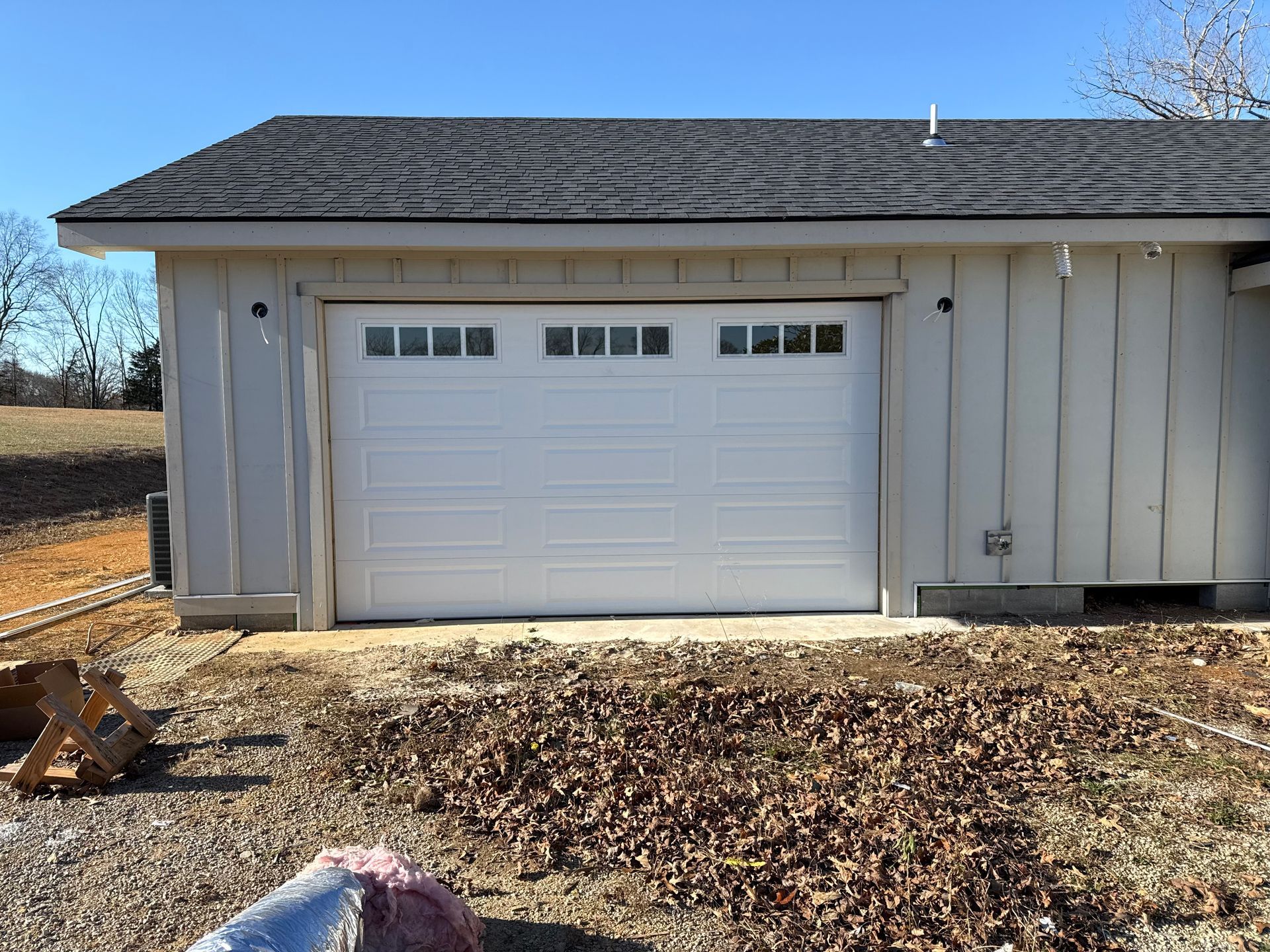 White garage door on a light-colored building under a dark roof, surrounded by brown leaves and dirt. White garage door on a light-colored building under a dark roof, surrounded by brown leaves and dirt.