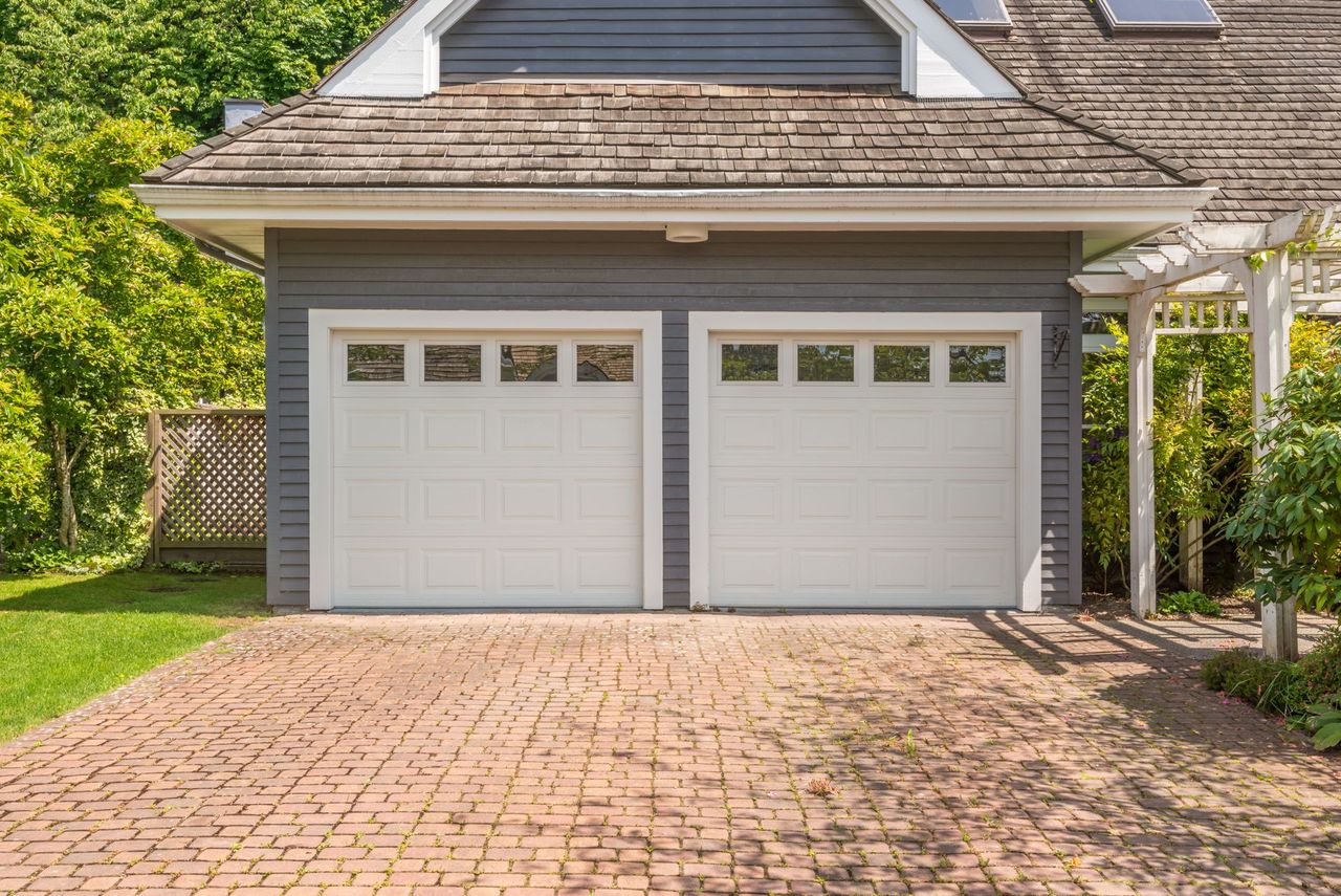 Two white garage doors with glass panels, gray exterior, brown brick driveway.