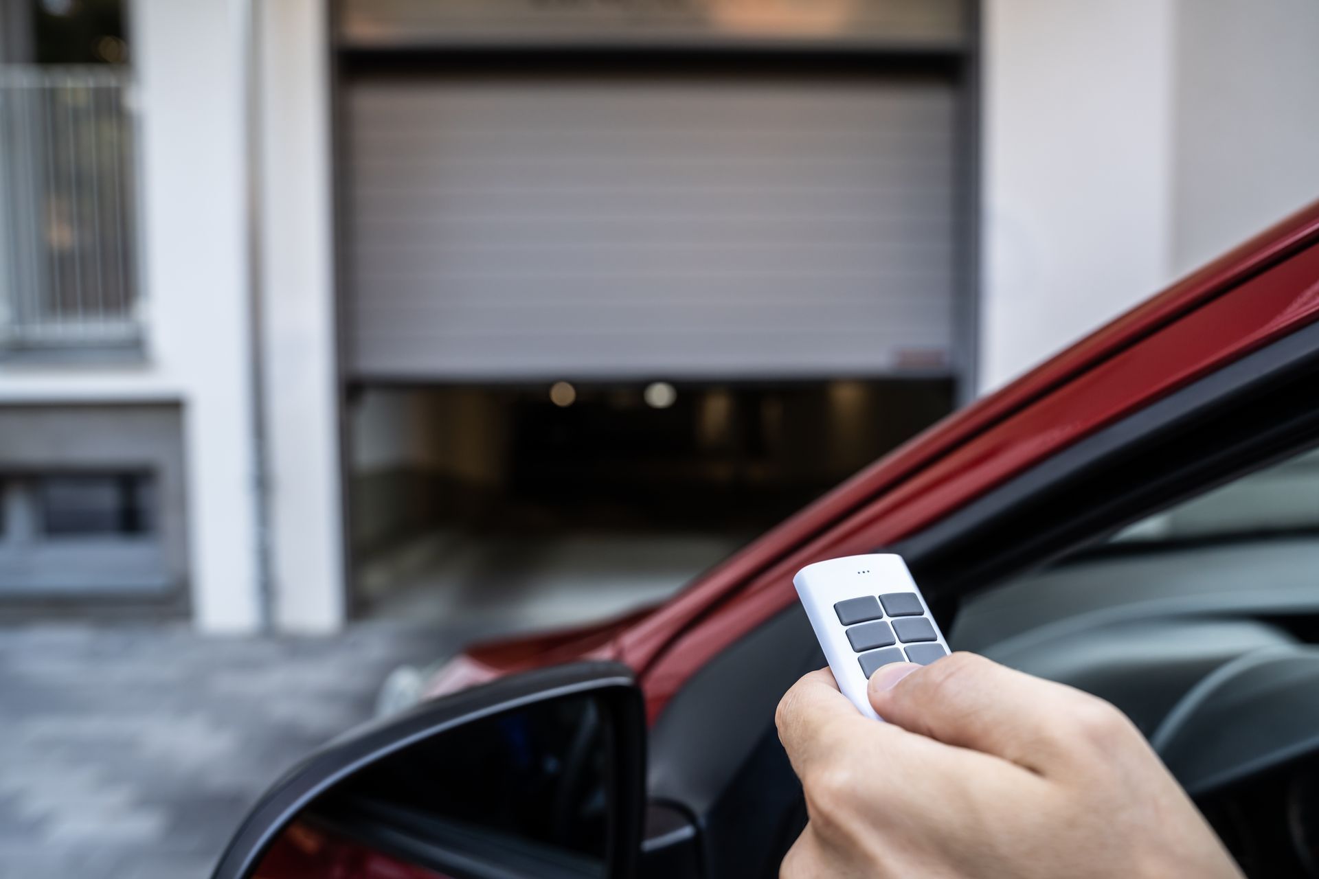 Hand holding a garage door opener near a red car, opening the garage door.