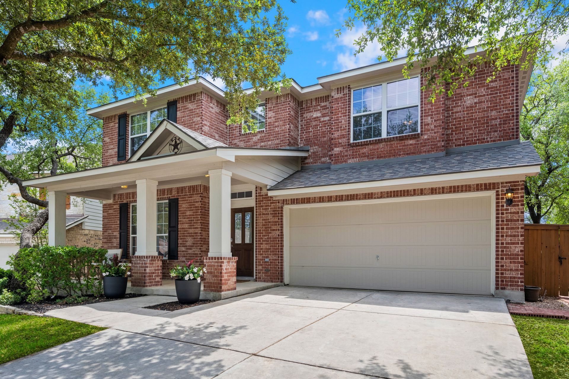 Two-story brick house with a porch and two-car garage under a blue sky, trees in the background.