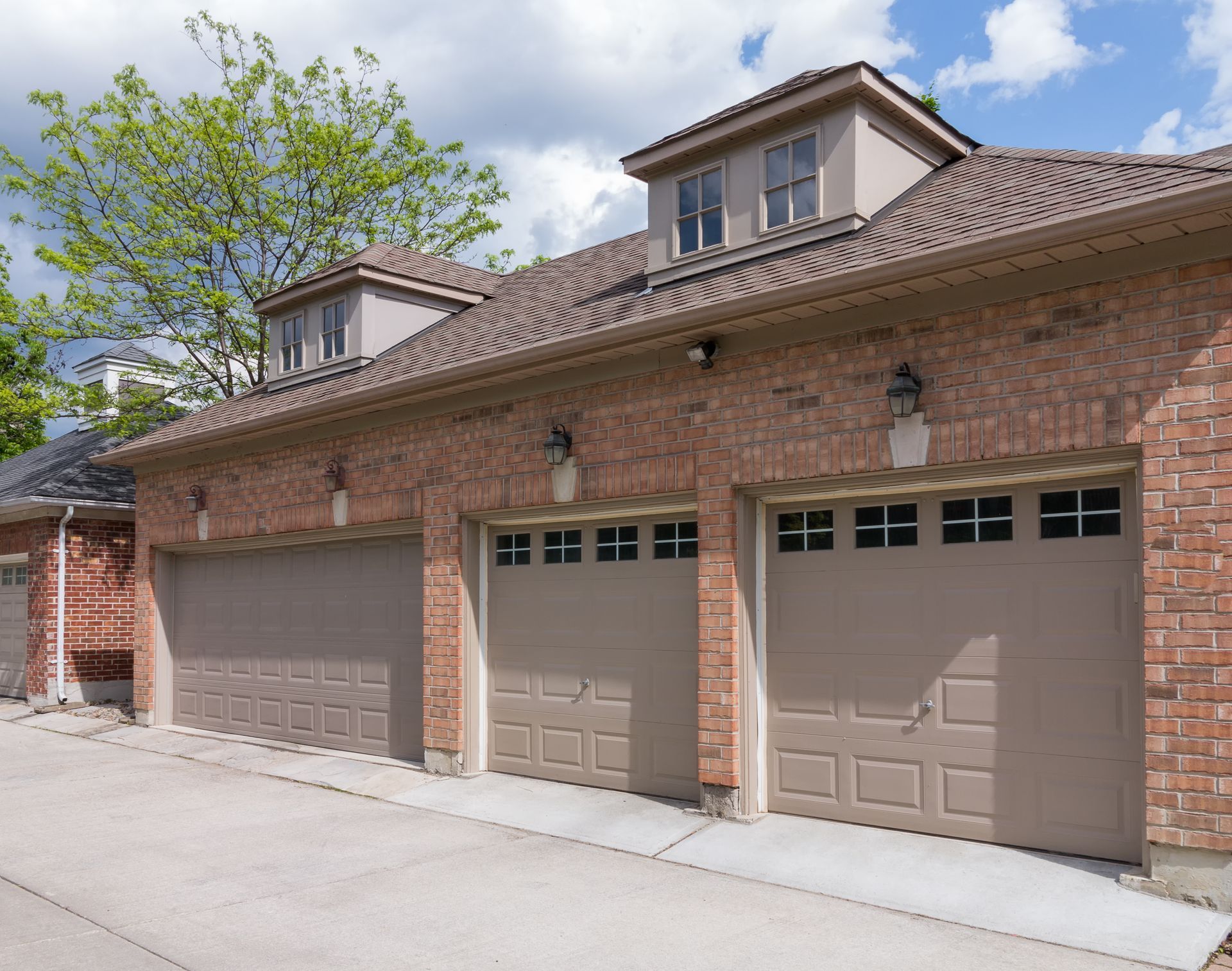 Three-car brick garage with tan doors, brown roof, and two dormers under a blue sky.