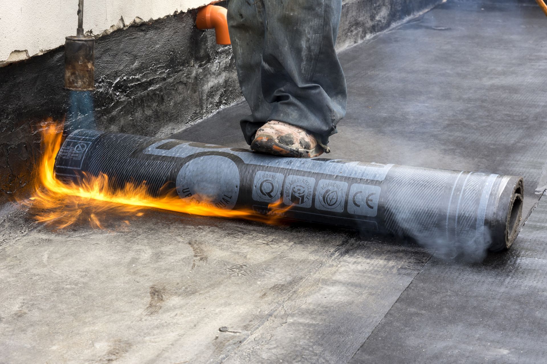 Roofer using a torch to install roofing material; flames visible, worker's foot on the roll.