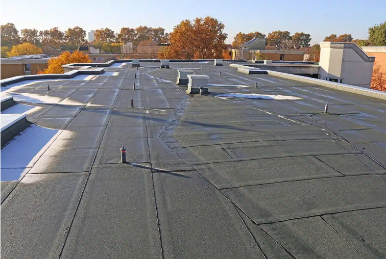 Flat black commercial roof with patches and vents, with autumn trees in the background.