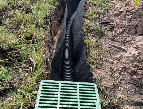 Drainage system installed in a trench. Black corrugated pipe inside fabric, leading to a green grate.