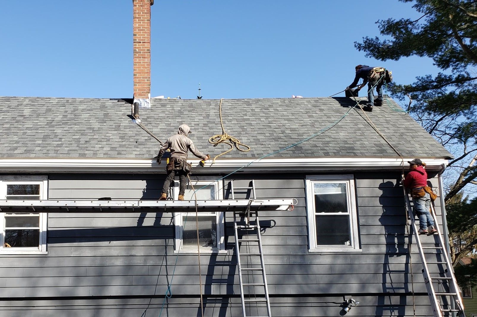 Three workers on a roof, installing shingles; ladder, chimney, gray siding and clear blue sky.