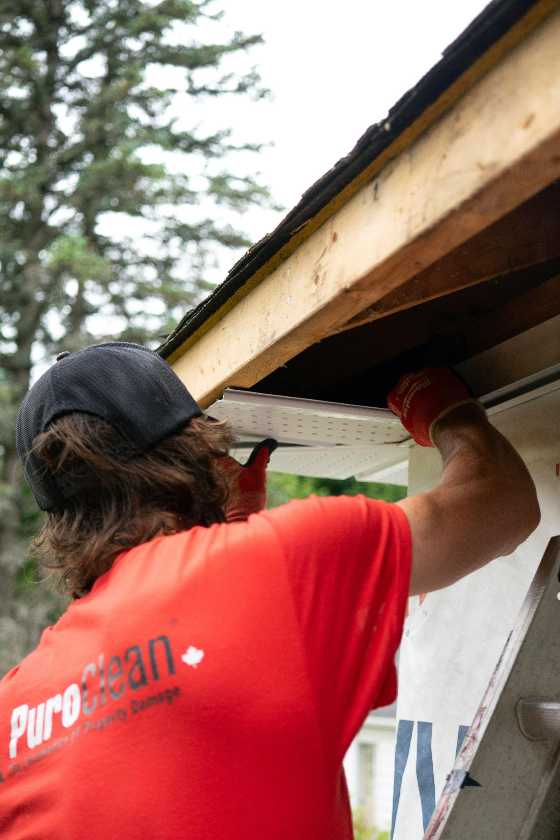 Person in red shirt installing white siding under a roof overhang.