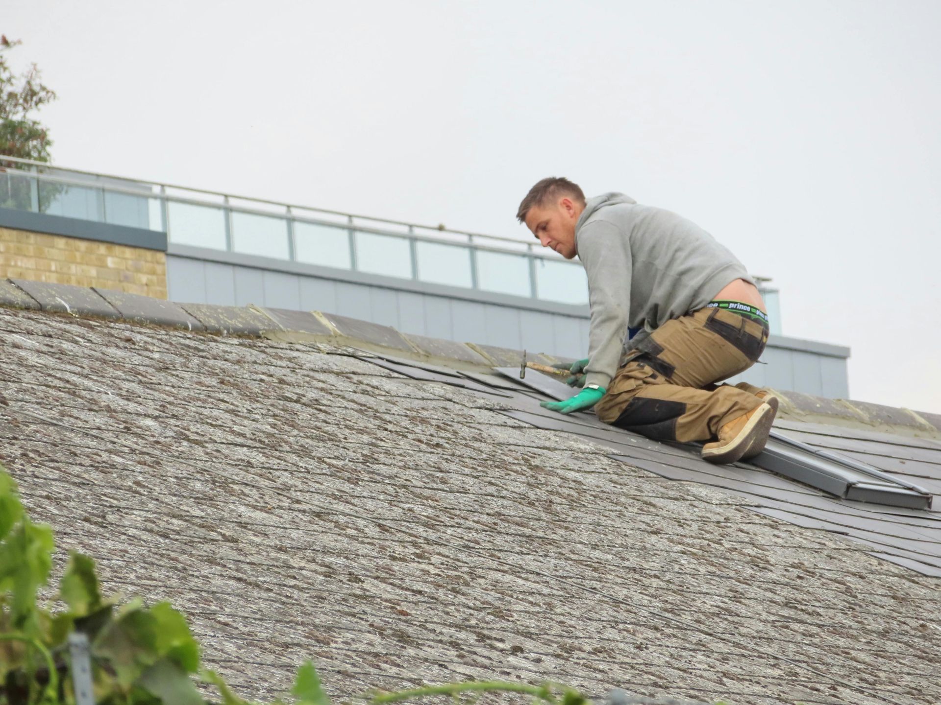 Roofer kneels on a weathered rooftop, working with a tool. Cloudy sky, building in background.