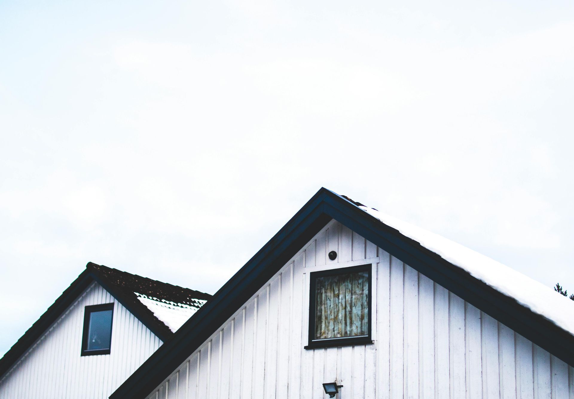 White house gabled roof with black trim and snow, against a pale blue sky.