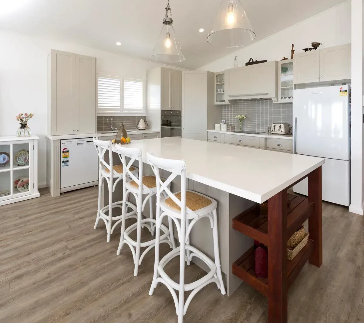 A modern kitchen with a large island, white cabinets, and wooden flooring; white bar stools.