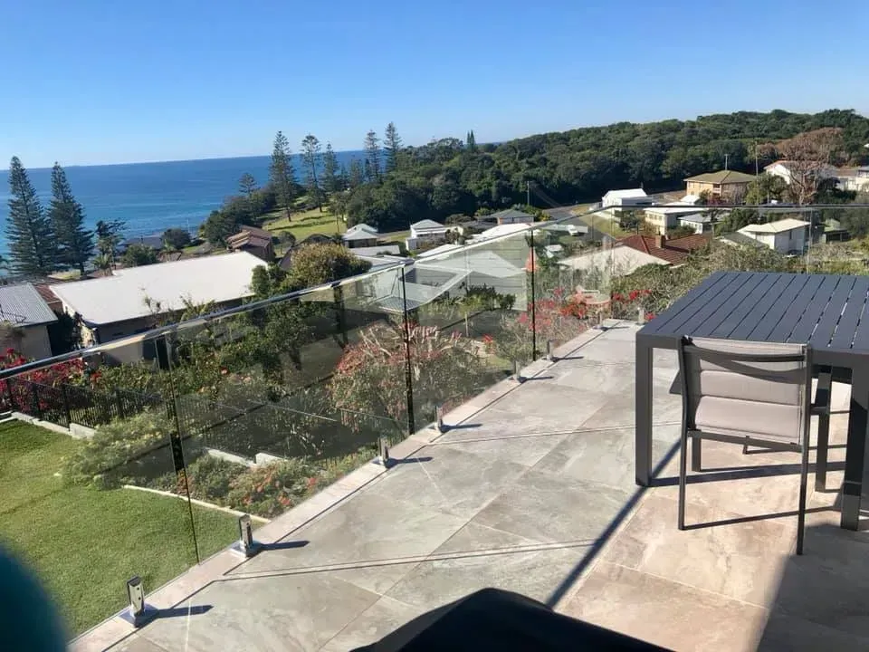 Coastal view from a patio: ocean, houses, table, chairs, glass railing, sunny day.