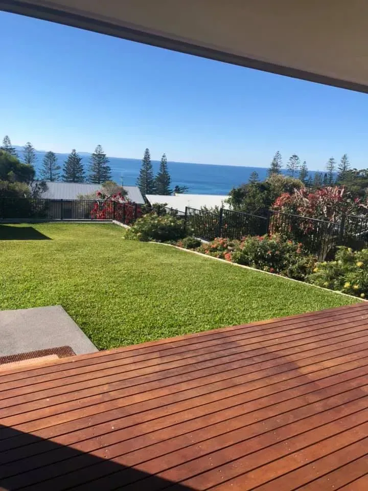 Wooden deck overlooking green lawn, lush garden, and ocean under a blue sky.