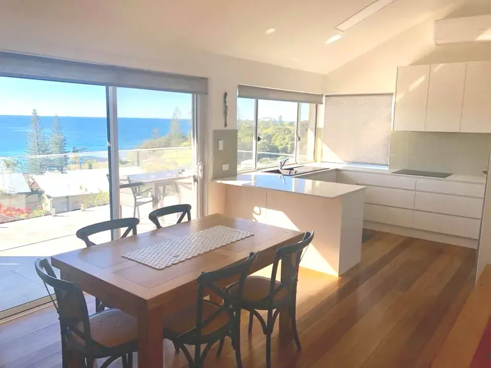Dining area with ocean view, wooden table and chairs, white kitchen, and large windows.